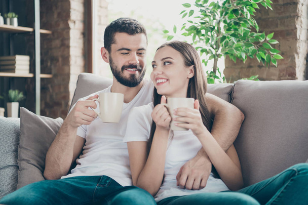 Photo of adorable pair in love holding hot beverage hands communicating sitting cosy sofa indoors - Fotografie, Obrázek