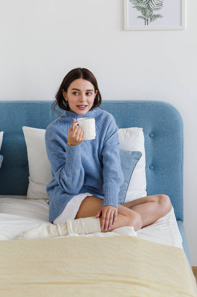  Girl in a blue sweater in hygge style interior with a cup of hot tea her hand sits on the bed - 写真・画像