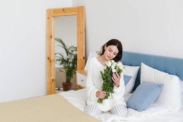 A girl in a white sweater sits on a bed holding a bouquet of white roses in her hand - Photo, image