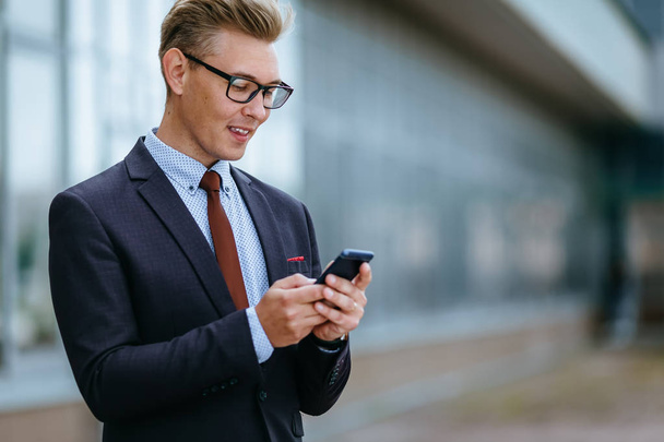 Happy businessman smiling as he reads a text message standing outside the office. Male executive manager in trendy clothes using smartphone outdoors. Modern technology concept - Photo, Image