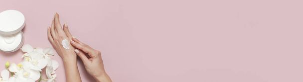 Jonge vrouw hydrateert haar hand met cosmetische crème lotion geopende container met crème lichaam melk witte Phalaenopsis orchidee bloemen op roze achtergrond plat leggen bovenaanzicht minimalisme stijl Beauty concept - Foto, afbeelding