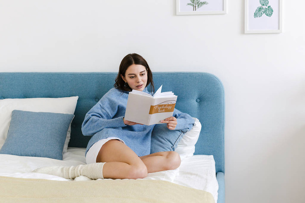 Girl in a blue sweater reading a book on psychology sitting on the bed in a cozy interior - Фото, зображення