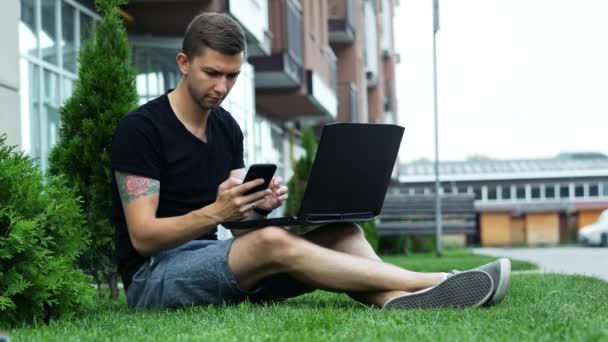 El hombre joven utiliza el teléfono móvil y el ordenador portátil mientras está sentado en la hierba
 - Metraje, vídeo