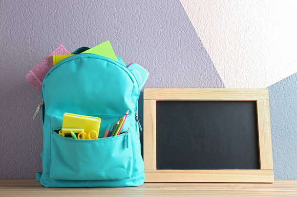 Backpack full of different school stationery and small blank chalkboard on table near color wall. Space for text - Foto, afbeelding