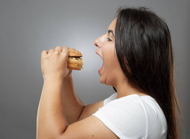 Woman eating hamburger, unhealthy fast food on gray background - Фото, изображение
