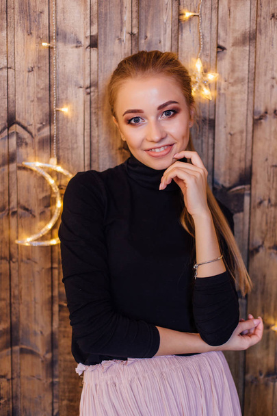 Beautiful young woman dressed in a sweater and skirt standing next to the wooden wall with star and moon lights - Fotografie, Obrázek