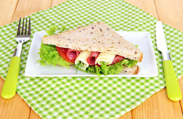 Tasty sandwich with salami sausage and vegetables on white plate, on wooden background - Valokuva, kuva