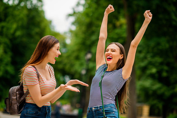 Portrait of delighted ladies with red lips stick screaming yeah raising fists closing eyes wearing striped t-shirt denim jeans bag backpack rucksack in city - Foto, imagen
