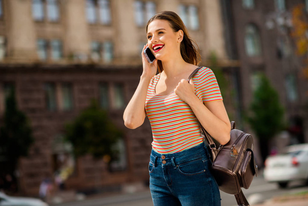 Profile side photo of cheerful lady with foxy ginger red lipstick hairstyle calling speaking wearing rucksack striped t-shirt denim jeans in city - Foto, Bild