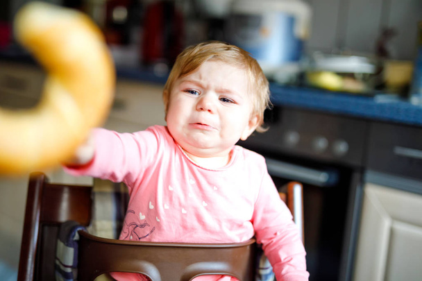 Милая маленькая девочка ест хлеб. Очаровательный ребенок ест в первый раз кусок кренделя или круассана. Здоровый плачущий ребенок. Первые зубы идут. Дома в помещении на кухне
. - Фото, изображение