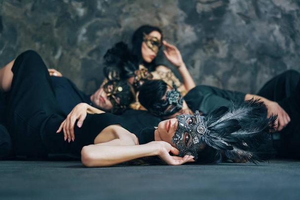 Group of friends in masquerade carnival masks laying on floor relaxing after party. Women and men wearing venetian masks. - Fotoğraf, Görsel