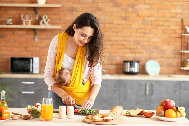 Young mother with little baby in sling preparing vegetable salad at home - Foto, Imagen