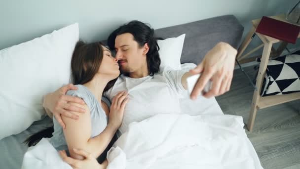 Husband and wife taking selfie with smartphone camera kissing hugging in bed - Кадри, відео