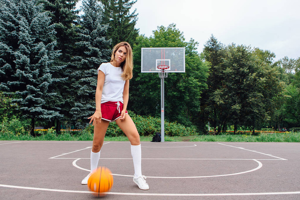 Beautiful young girl dressed in white t-shirt, shorts and sneakers, plays with a ball on a basketball court. - 写真・画像