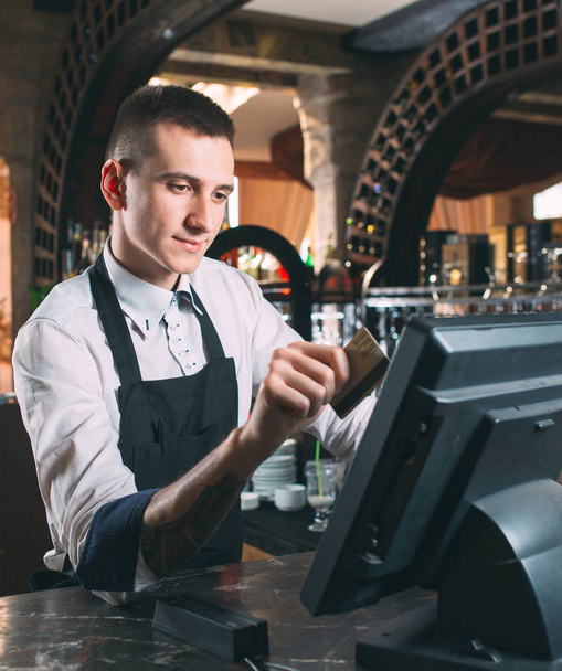 small business, people and service concept - happy man or waiter in apron at counter with cashbox working at bar or coffee shop - Фото, зображення