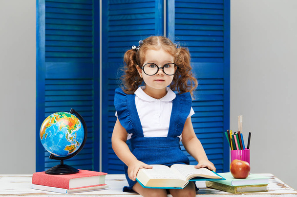 Smart toddler kid. Back to school and happy time! Cute industrious child in glasses is sitting at a desk indoors. Kid is learning in class on background of blue wall. Girl reading the book. - Photo, Image