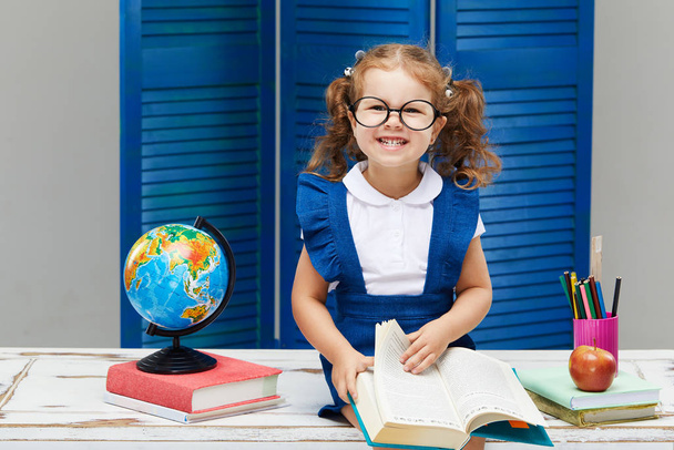 Smart toddler kid. Back to school and happy time! Cute industrious child in glasses is sitting at a desk indoors. Kid is learning in class on background of blue wall. Girl reading the book. - Fotó, kép