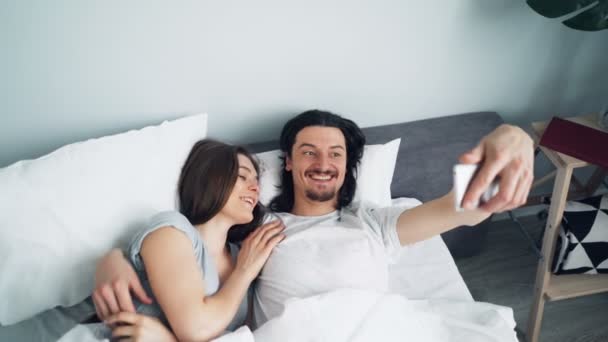 Girl and guy taking selfie in bed with smartphone camera kissing hugging - Metraje, vídeo