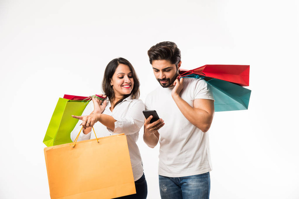 Indiai fiatal pár bevásárló táskák és okos telefon vagy mobil, kattintson selfie vagy helyüket tárolni, állandó izolált fehér háttér előtt. szelektív összpontosítás - Fotó, kép
