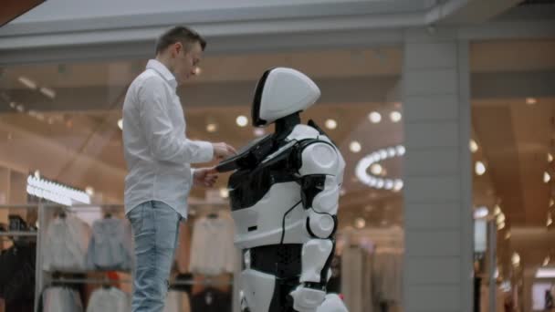 Un hombre con camisa se comunica con un robot blanco haciendo preguntas y presionando la pantalla con los dedos
. - Metraje, vídeo