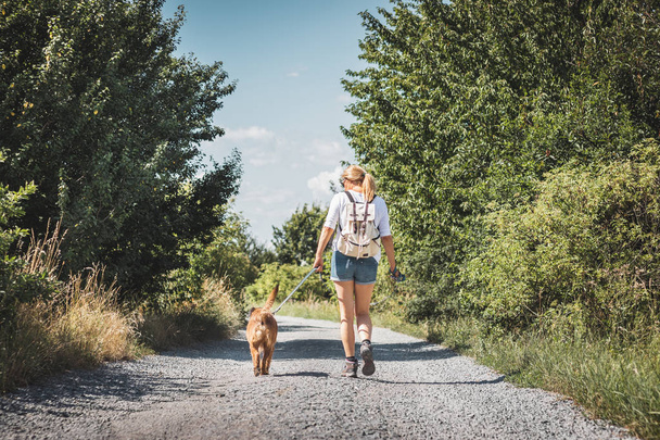 Frau und Hund gehen gemeinsam auf Feldweg. Touristin mit Rucksack genießt Spaziergang mit ihrem Tier beste Freundin - Foto, Bild