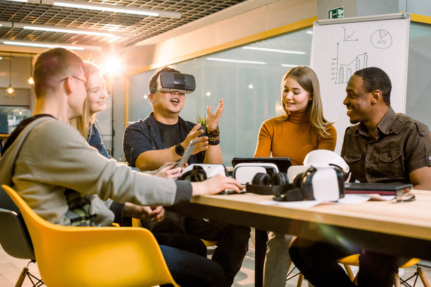 Teamwork trying virtual reality glasses for work in creative office. Young Asian man using new technology vr goggles while his coworkers talking sitting at the table - Photo, Image
