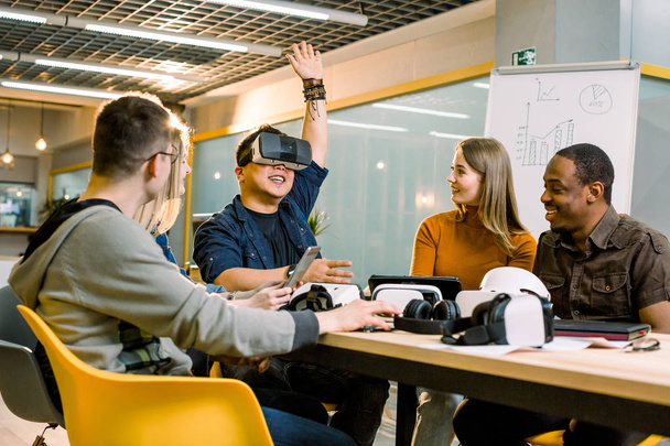 Five young funny people sitting at the table in front of each other, one man is using virtual reality goggles. VR goggles concept, business meeting - Zdjęcie, obraz