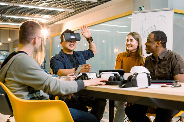 Five young funny people sitting at the table in front of each other, one man is using virtual reality goggles. VR goggles concept, business meeting - Photo, Image