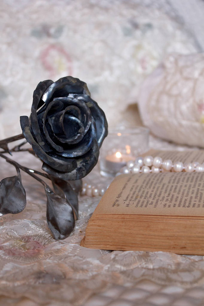 Hygge-Stil. Eine brennende Kerze erhellt ein aufgeschlagenes altes Buch. Auf dem Buch liegt eine Perlenkette. In der Nähe liegt eine geschmiedete Rose als Symbol der ewigen Liebe. Vintage-Stil, Stillleben. - Foto, Bild