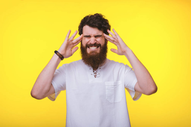 Un hombre barbudo está mostrando que tiene un dolor de cabeza cerca de una pared amarilla
 - Foto, imagen