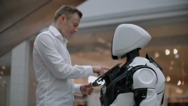 Un uomo sta con un robot robot e gli fa domande e chiede aiuto facendo clic sullo schermo sul corpo del robot
. - Filmati, video