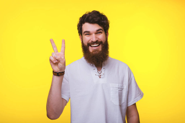 Cheerful young bearded man is showing peace sign on a yellow background. - Photo, Image