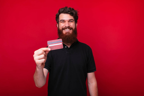 Χαρούμενος νέος άνθρωπος με γένια δείχνει μια πιστωτική κάρτα σε ένα κόκκινο φόντο. - Φωτογραφία, εικόνα