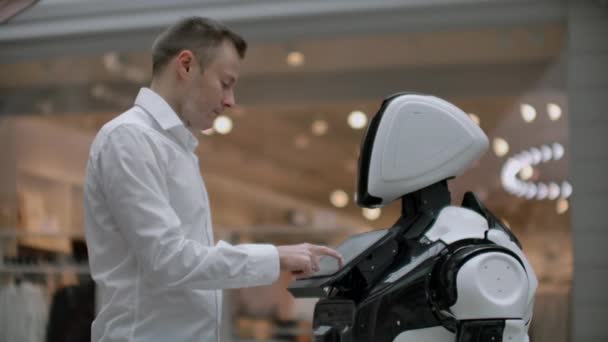 A man in a shirt communicates with a white robot asking questions and pressing the screen with his fingers. - Footage, Video