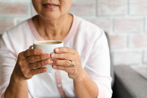 Old woman hold a cup of hot coffee drink at home - Foto, Imagen