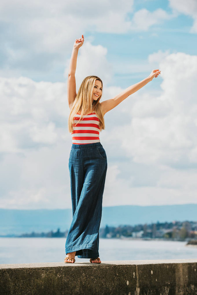Outdoor fashion portrait of beautiful young woman posing by the lake, wearing red and white stripe vest and denim wide leg jean - Photo, Image
