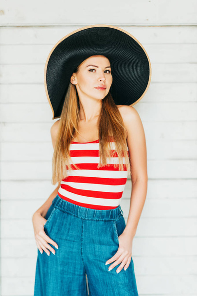 Outdoor fashion portrait of beautiful young woman posing on white background, wearing red and white stripe vest, denim wide leg jean and big black hat - Zdjęcie, obraz