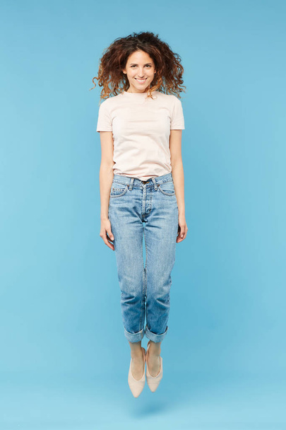 Счастливая молодая случайная женщина в джинсах и футболке прыгает через синий фон в изоляции перед камерой
 - Фото, изображение