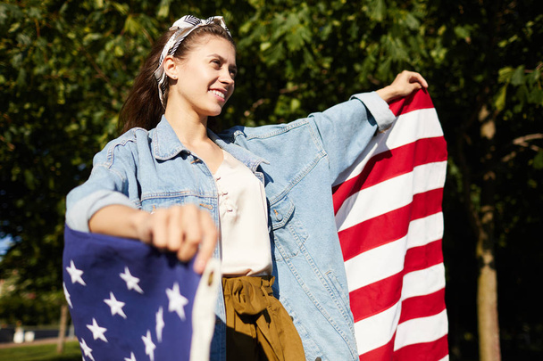 Cheerful pretty young woman in long denim jacket standing in summer forest and holding big American flag while celebrating independence day - Photo, Image