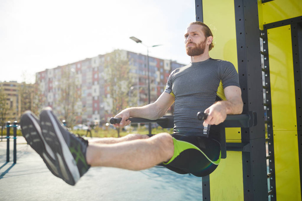 Concentrated young bearded man in sportswear hanging on parallel bars and exercising on bars outdoors - Photo, Image