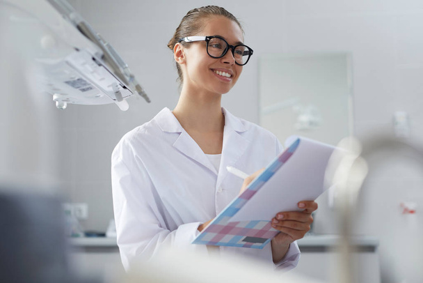 Portrait d'une jeune dentiste tenant un presse-papiers et souriant joyeusement tout en consultant la patiente, espace de copie
 - Photo, image