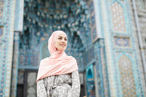 Contenido atractiva joven musulmana en hijab de pie contra la colorida mezquita con adornos y mirando hacia otro lado
 - Foto, Imagen