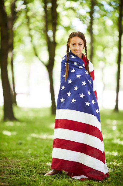 zufriedenes hübsches Mädchen mit Zöpfen in amerikanische Flagge gehüllt, das im Sommerpark steht und in die Kamera blickt - Foto, Bild