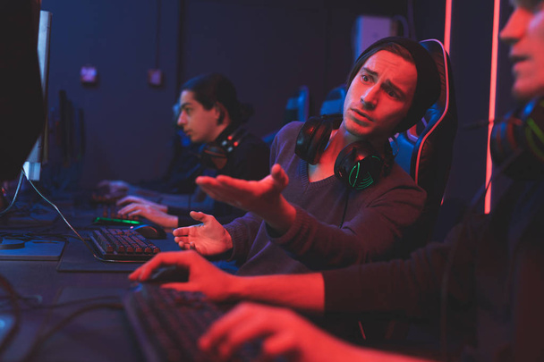 Hipster teen in beanie wearing headset on neck sitting at table and pointing with hand at monitor while giving advice to friend about computer game - Foto, Imagen