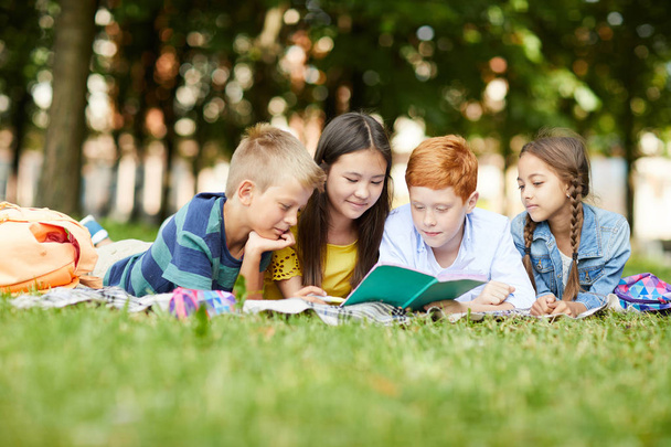 Cuatro amigos adolescentes de la escuela leyendo juntos historia para la clase de literatura en el parque acostado en la hierba después de clases en la escuela
 - Foto, Imagen