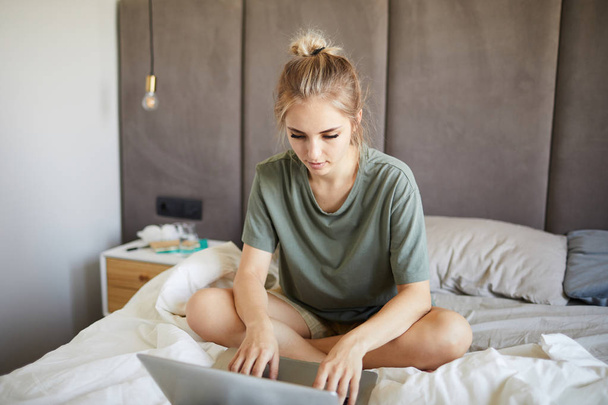 Linda chica rubia en camiseta escribiendo y mirando la pantalla del ordenador portátil mientras está sentado en la cama por la mañana
 - Foto, imagen