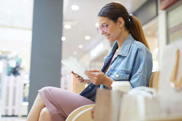 Jong glimlachend aantrekkelijk meisje zitten en lezen van Tablet in winkelcentrum terwijl je na het winkelen breekt - Foto, afbeelding