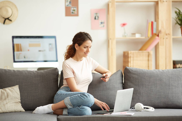 Веселая современная девушка в футболке и джинсах сидит дома на диване и использует дебетовую карту при покупке товаров в интернет-магазине
 - Фото, изображение