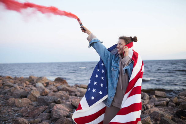 Cheerful excited young woman in casual outfit covered with American flag standing on rocky shore and waving colored smoke bomb on Independence day - Photo, Image