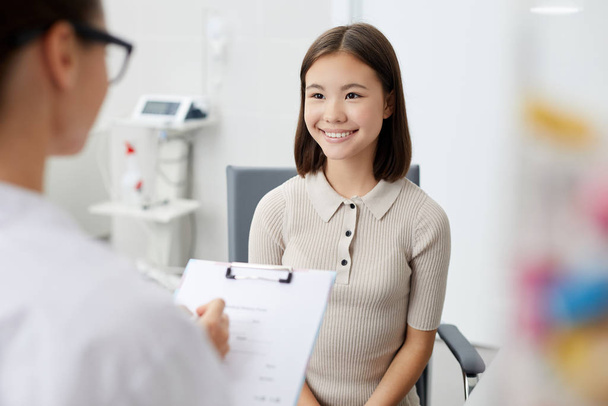 Retrato de menina asiática olhando para o médico e sorrindo feliz durante a consulta na clínica, espaço de cópia
 - Foto, Imagem